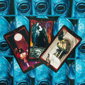 Tarot kortos gotikiniu raštu ir mėlynomis nugarėlėmis, trys atverstos kortos su raudonais rėmeliais ir auksinėmis taurėmis centre, pateiktos kampuotu išdėstymu