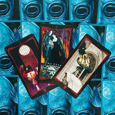 Tarot kortos gotikiniu raštu ir mėlynomis nugarėlėmis, trys atverstos kortos su raudonais rėmeliais ir auksinėmis taurėmis centre, pateiktos kampuotu išdėstymu