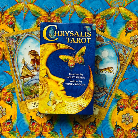 Chrysalis Tarot kortų rinkinys su ryškiai mėlyna dėže ir geltonomis drugelių iliustracijomis, dėžė centre ant išskleistų kortų fono