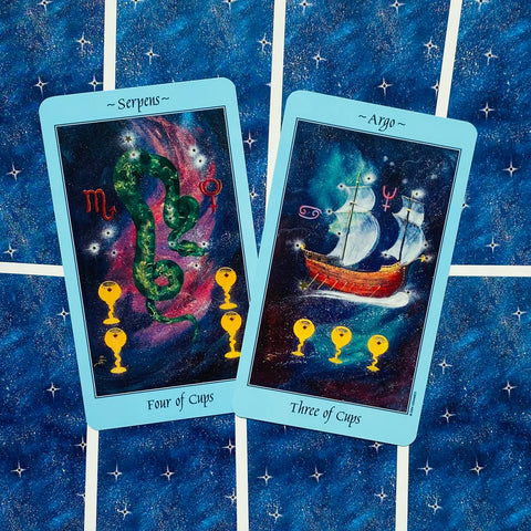 tarot kortos mėlynu rėmeliu, dvi atverstos kortos (Four of Cups ir Three of Cups) ant žvaigždėto mėlyno audinio