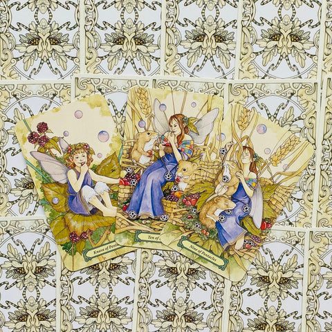 Taro kortos su spalvotomis fėjų iliustracijomis — trys atvertos kortos centre ir ornamentinis nugarėlių raštas