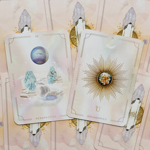 Orakulų kortos su pasteliniais rožiniais atvartais, dvi atverstos — Larimar (Perspective) ir Labradorite (Boundaries), fone krištolai