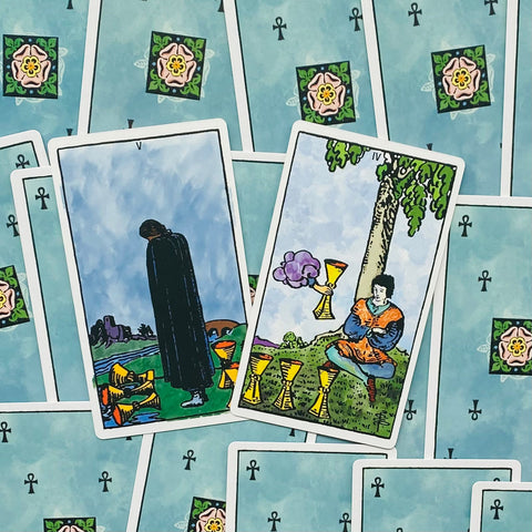 Tarot kortos Rider–Waite stiliaus, mėlynos nugarėlės su rozetėmis, parodytos dvi priekinės kortos: Keturi taurės ir Penki taurės