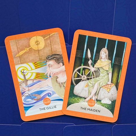 Tarot kortos su oranžinėmis kraštinėmis, dvi atviros kortos (The Gillie, The Maiden) iš viršaus ant mėlyno fono, matomos iliustracijos
