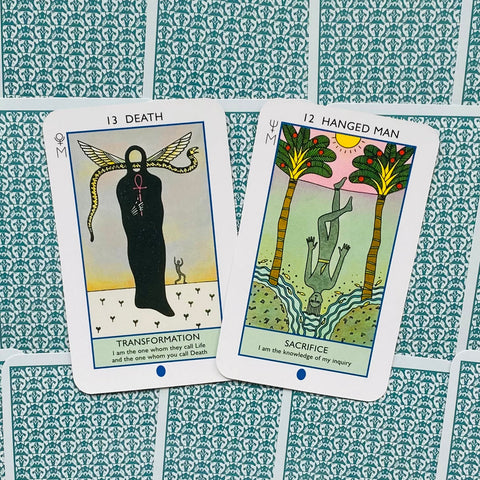 Tarot kortos, dvi atverstos kortos Death ir Hanged Man su ryškiais piešiniais ant žalsvai raštuotų nugarėlių, matomas priekinis kortelių paviršius