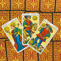Tarot kortos Marseilio stiliaus — trys Pentaklių kortos (karalienė, riteris, jaunuolis) išsidėsčiusios ventyla ant raudono‑geltono raštuoto nugarėlių fono