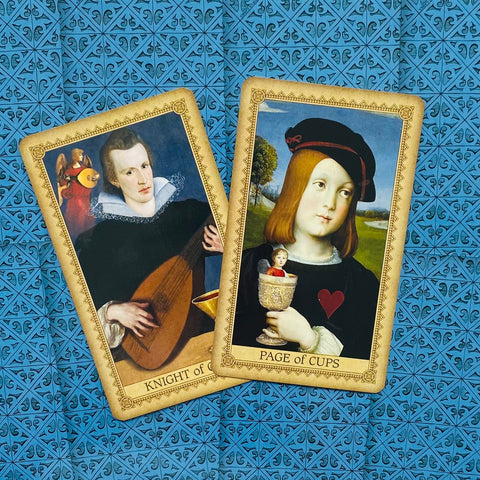 Tarot kortos su Renesanso paveikslų dizainu, dvi kortos (Knight of Cups ir Page of Cups) ant mėlyno raštuoto fono, fotografuotos iš viršaus