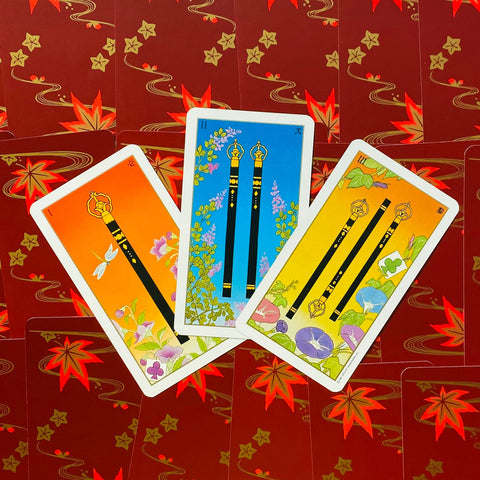 Tarot kortos rinkinys, trys atvertos lazdelės kortos su oranžinėmis, mėlynomis ir geltonomis priekinių pusių detalėmis ant raudono fono