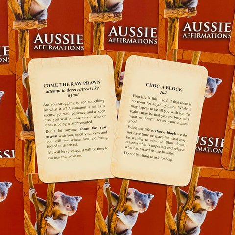Aussie Affirmations kortos atviros dviem kortomis — Come the Raw Prawn ir Choc‑a‑Block — ant raudono koalos fono, matomas kortų tekstas