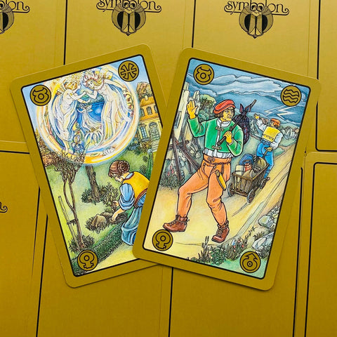 tarot kortos Symbion, aukso rėmeliai ir detalūs iliustracijos, dvi kortos padėtos išskėstai ant geltono dėklo, unikalios variantų iliustracijos