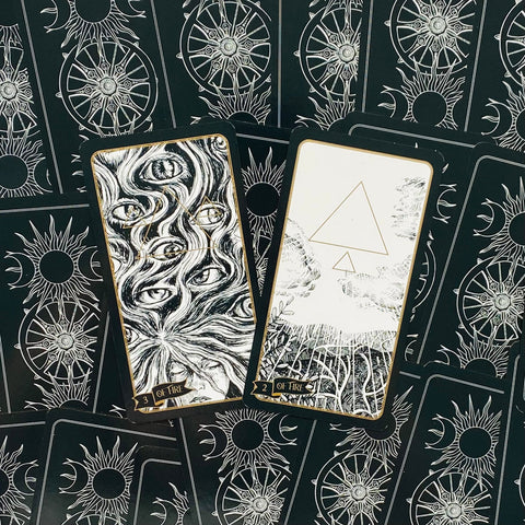tarot kortos juodai baltos su auksiniu apvadu, dvi atverstos kortos (akių motyvas ir trikampis) ant nugarėlių su saulėmis ir ratais