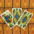 Tarot kortos Marseille stiliaus, ryškios geltonos–mėlynos lazdelės ant trijų atverstų kortų prie raudono‑auksinio nugarėlių rašto