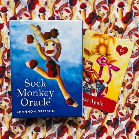 Orakulo kortų rinkinys Sock Monkey Oracle mėlyname dėkle su iliustruotu kojinių beždžionės piešiniu ir atversta 32 korta ant raštuoto fono