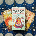 Tarot kortos Gregory Scott dėžutėje su baltu viršeliu, centre ant mėlynų žvaigždžių nugarėlių, trys atverstos kortos rodytos fanuotos