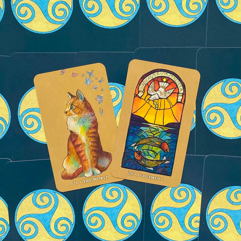 tarot kortos mėlynoje nugarėlėje, centre „The World“ su katės iliustracija ir „Judgement“ su vitražiniu unguriu ir balandžiu