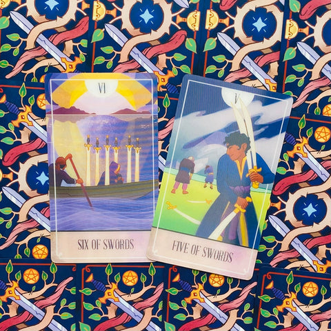 Tarot kortos rinkinys — dvi atviros kortos (Six of Swords ir Five of Swords) rodomos priekyje ant ryškaus kardų rašto fono, unikalus variantas