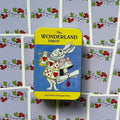 Produktų puslapyje: geltona The Wonderland Tarot skardinė su baltuoju triušiu, centre ant išklotų violetinių kortų nugarėlių, matomas priekinis tin variantas.