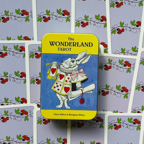 Produktų puslapyje: geltona The Wonderland Tarot skardinė su baltuoju triušiu, centre ant išklotų violetinių kortų nugarėlių, matomas priekinis tin variantas.