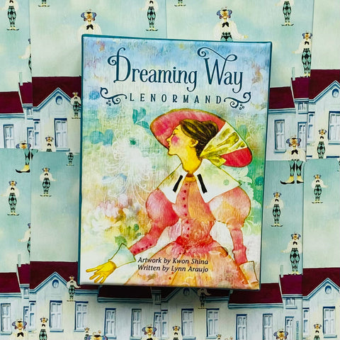 Dreaming Way Lenormand kortų rinkinys su spalvota moters iliustracija ant priekinės dėžutės, rodoma iš viršaus ant pastelinio raštuoto fono