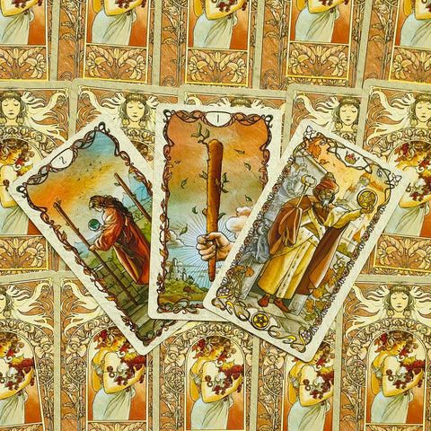 Tarot kortos art nouveau stiliaus, šiltos okerio ir aukso spalvos, trys išskleistos kortos su Lazdos tūriu centre, detali ornamentika