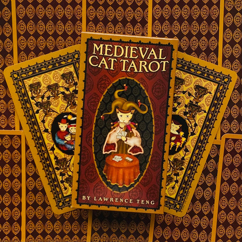 Medieval Cat Tarot stalo kortų rinkinys (Lawrence Teng) su raudona–auksine dėže ir dviem išskleistomis nugarėlėmis, skirtas taro praktikoms ir kolekcijai