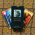 Tarot kortos Tarot of the Elves juoda dėžutė centre su dviem spalvingomis elfų iliustracijų kortomis