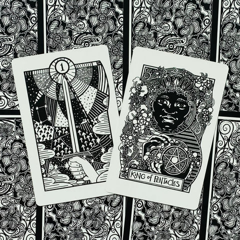 Tarot kortos juodai baltos iliustruotos, Karalius pentaklių ir Asas kardas ant gėlių nugarėlių, išdėstytos ant stalo