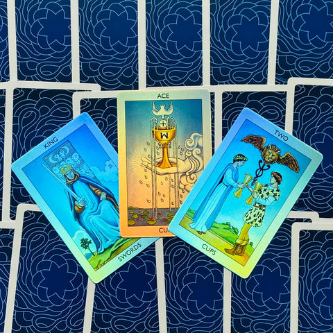 Tarot kortos Rider–Waite stiliaus su tamsiai mėlynomis nugarėlėmis, trijų kortų išdėstymas atverstas (Ace of Cups, King of Swords, Two of Cups), rodantis kortų priekį ir detales