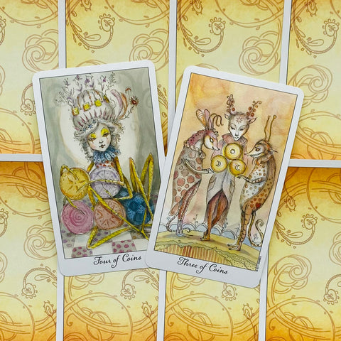 Tarot kortos su akvarelinėmis iliustracijomis — dvi atviros monetų kortos (Three/Four of Coins) prieš geltonas ornamentuotas nugarėles, skirtos komplekto parodymui