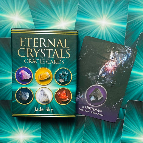 Oracle kortų rinkinys Eternal Crystals (Jade‑Sky) žalsvame dėkle, šalia atversta Obsidian korta nurodanti apsaugą ir šaknų čakrą