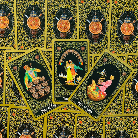 Tarot kortos juodu fonu su auksiniu ornamentu, trys atverstos kortos centre (Nine of Cups, Ten of Cups, Page of Cups)