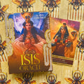 Isis Oracle kortos Alana Fairchild dėžutėje su auksuotais akcentais ir atversta viena korta šone, rodanti meno variantą