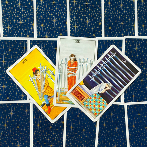 Tarot kortos su tamsiai mėlynomis nugarėlėmis su žvaigždžių raštu ir trimis kardų kortomis (VII–IX) išskleistos ant stalo