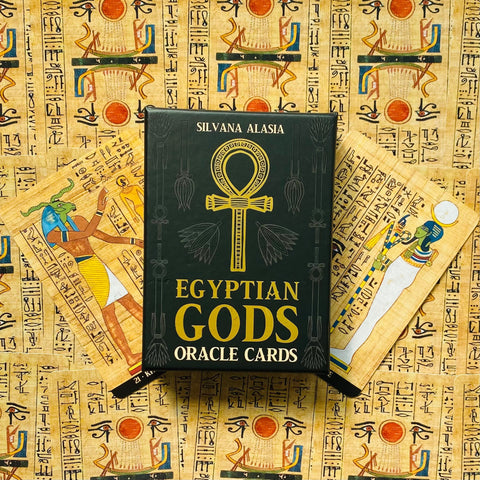 Egyptian Gods orakulo kortos — juoda auksuota dėžutė iš priekio, dvi papiruso stiliaus kortos išskleistos šonuose