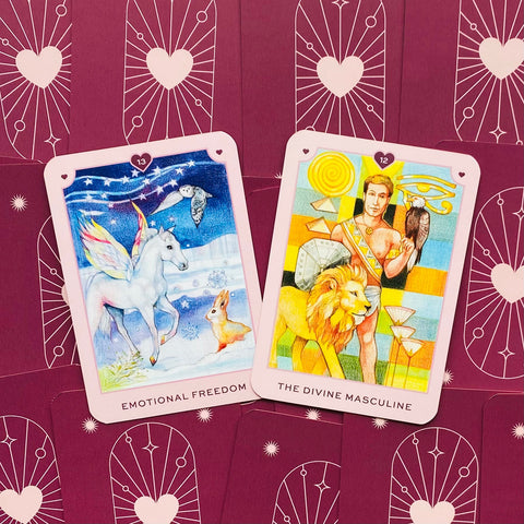 tarot kortos rožiniais rėmeliais — dvi atviros kortos (Emotional Freedom ir The Divine Masculine) ant bordo fono, aiškios iliustracijos
