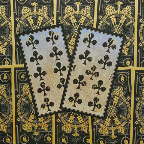 Tarot kortos su juoda ir aukso nugarėle, dvi atverstos klubų kortos pasuktos kampu ant ornamentuoto fono