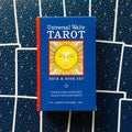 Universal Waite Tarot dėžutė su knyga, mėlynas viršelis ir geltonas saulės paveikslas, raudonas šonas, nufotografuota prieš žvaigždėtą kortelių foną