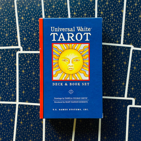Universal Waite Tarot dėžutė su knyga, mėlynas viršelis ir geltonas saulės paveikslas, raudonas šonas, nufotografuota prieš žvaigždėtą kortelių foną