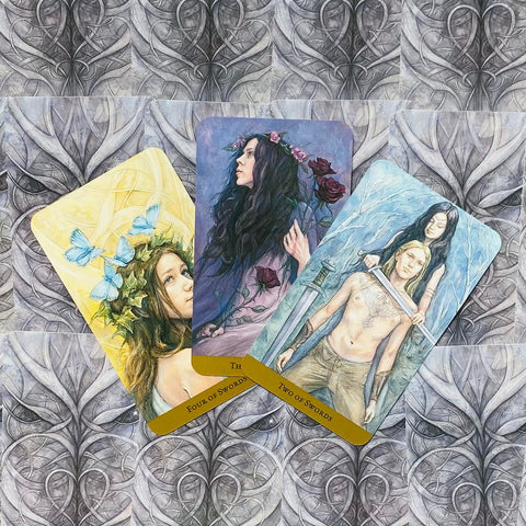 Tarot kortos trijų kortų išdėstymas: centrinė profilinė moteris su rožėmis, dešinėje vyriškis ir moteris su kardais, kairėje geltona korta su drugeliais