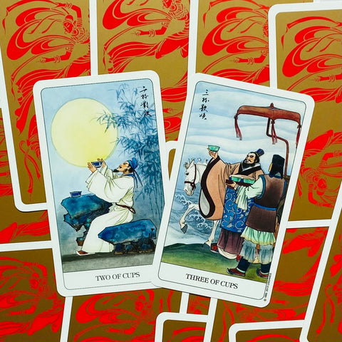 Tarot kortos su kinų stiliaus iliustracijomis — atverstos Two of Cups ir Three of Cups, artimas kampas ir raudonai auksinės nugarėlės