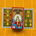 Oswald Wirth tarot kortos: raudonu rėmu pakuotės priekinė dalis centre tarp dviejų taurės kortų ant geltono raštuoto fono, išryškinta dėžutės iliustracija