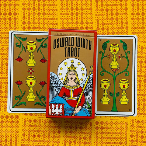 Oswald Wirth tarot kortos: raudonu rėmu pakuotės priekinė dalis centre tarp dviejų taurės kortų ant geltono raštuoto fono, išryškinta dėžutės iliustracija