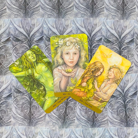 Tarot kortos – trijų kortų rinkinys su geltonai‑žaliomis iliustracijomis, centre mergaitė laiko sėklą ant ornamentuoto fono