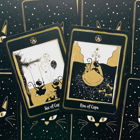Tarot kortos juodos su auksinėmis detalėmis ir katinų iliustracijomis, dvi atidengtos kortos (Five ir Six of Cups) ant žvaigždėto fono