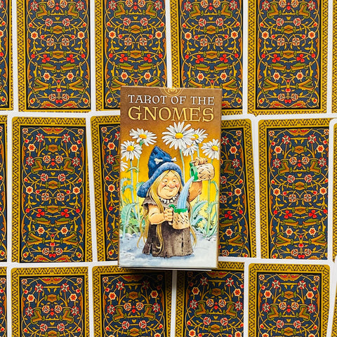 Produktų puslapyje Tarot of the Gnomes dėžutė centre, gnomis pilsto vandenį prie baltų ramunių ant dekoratyvių kortų nugarėlių