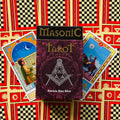 Masoniškas tarot rinkinys su bordo dėkle, dviem pavyzdiniais kortomis ir ryškia raudono‑geltono langų fono kompozicija