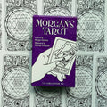 Tarot kortos Morgan's Tarot violetinėje dėžutėje, priekyje ranka laiko kelias kortas, fone juodai baltų nugarėlių raštai