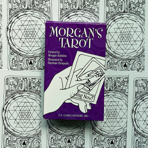 Tarot kortos Morgan's Tarot violetinėje dėžutėje, priekyje ranka laiko kelias kortas, fone juodai baltų nugarėlių raštai