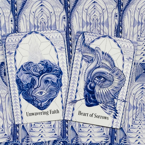 Kortos su mėlynomis iliustracijomis, artimas dviejų kortų komplekto priekinis kampas, kuriame 'Unwavering Faith' ir 'Heart of Sorrows'