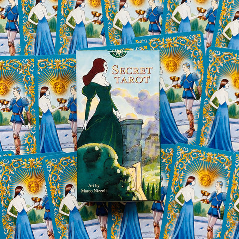 Secret Tarot kortų rinkinys su Marco Nizzoli iliustracijomis, žalsvai mėlyna dėžutė centre ant išklotų aukso‑mėlynų kortų fono